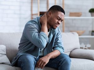 Man sitting on a sofa holding his neck, appearing to experience neck pain or discomfort.