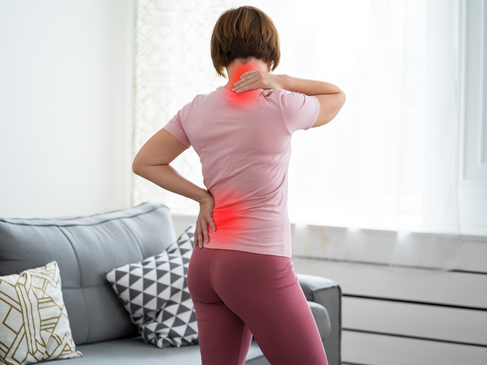 Woman standing indoors holding her neck and lower back, with highlighted areas indicating neck and back pain.